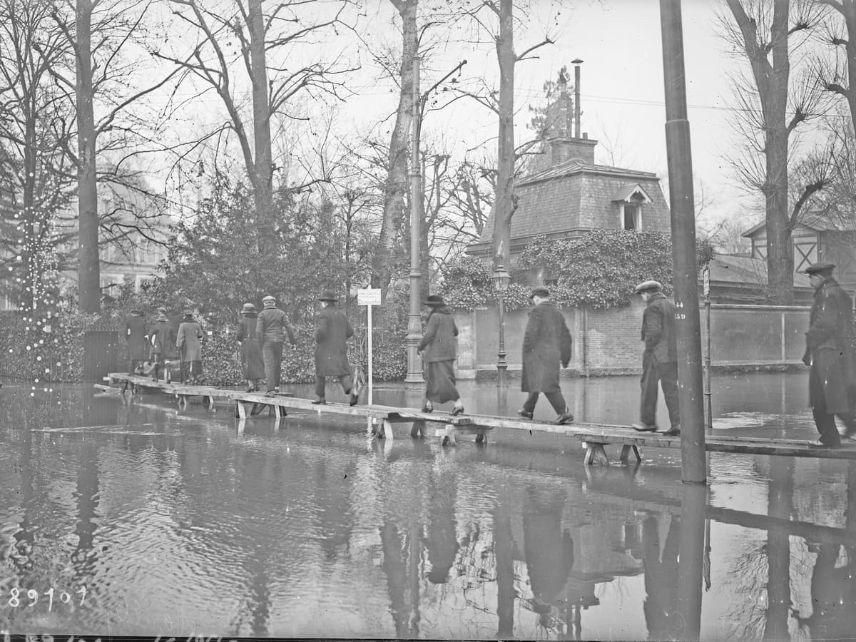 Photographie de 1924 : des passants avancent sur une passerelle de planches au-dessus d'une rue inondée, entre des arbres nus.
